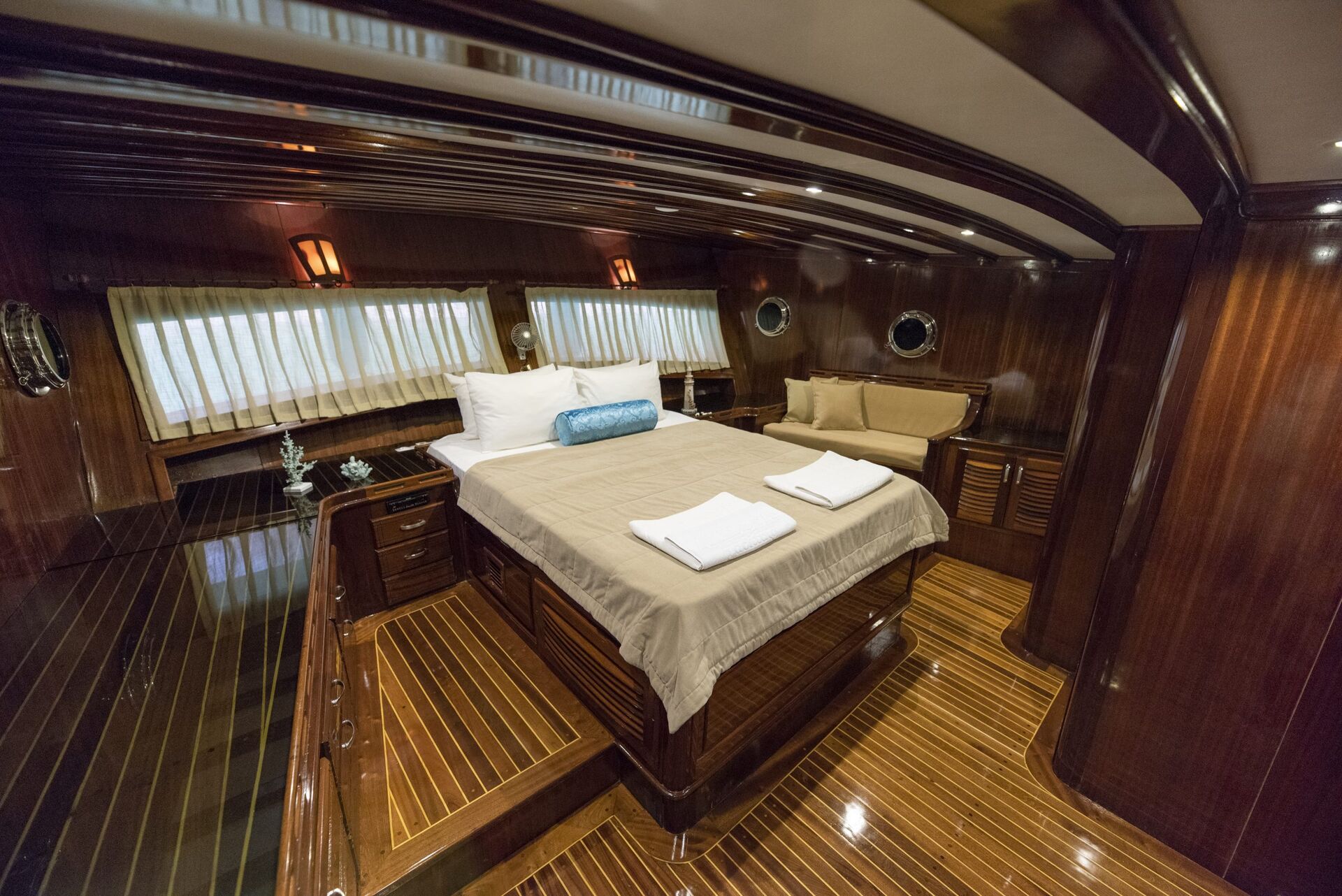 Luxurious yacht cabin with polished wood walls and floor, a large bed with beige bedding, white towels, decorative pillows, small windows with curtains, and warm ambient lighting.