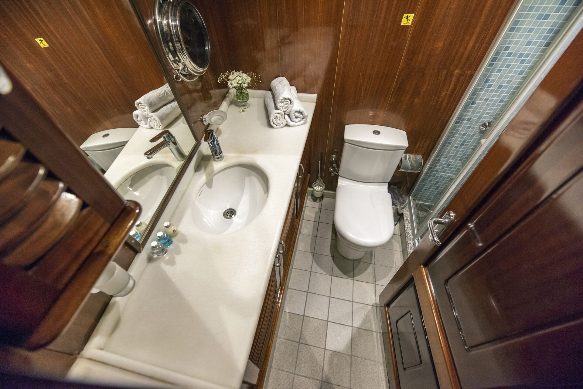 A compact, modern bathroom with wood-paneled walls, a white sink and countertop, a toilet, a glass shower, a mirror, and neatly arranged towels and toiletries.