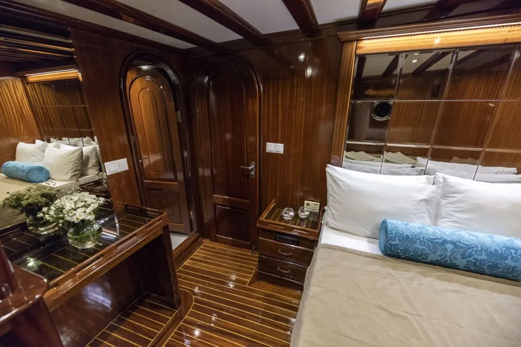 Luxurious yacht cabin with glossy wooden walls and floor, a neatly made bed with white pillows and blue cushions, a mirrored wall, bedside table, flowers, and a doorway leading to another room.