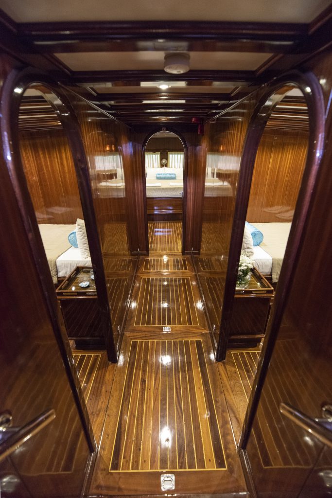 Polished wooden hallway inside a yacht leads to a cozy cabin with two single beds, soft lighting, and elegant decor reflecting on the shiny floor.