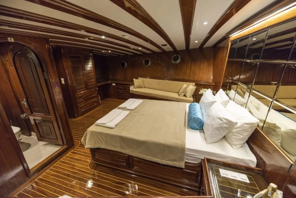 Luxurious yacht bedroom with polished wood walls and floor, a large bed with white and beige linens, a beige sofa, small porthole windows, mirrors, and a doorway leading to a bathroom.
