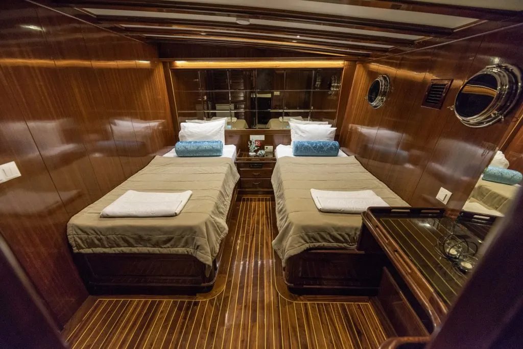 Luxurious yacht cabin with two single beds, beige bedding, blue accent pillows, wood-paneled walls, glossy wooden floor, porthole windows, and a nightstand with a telephone between the beds.