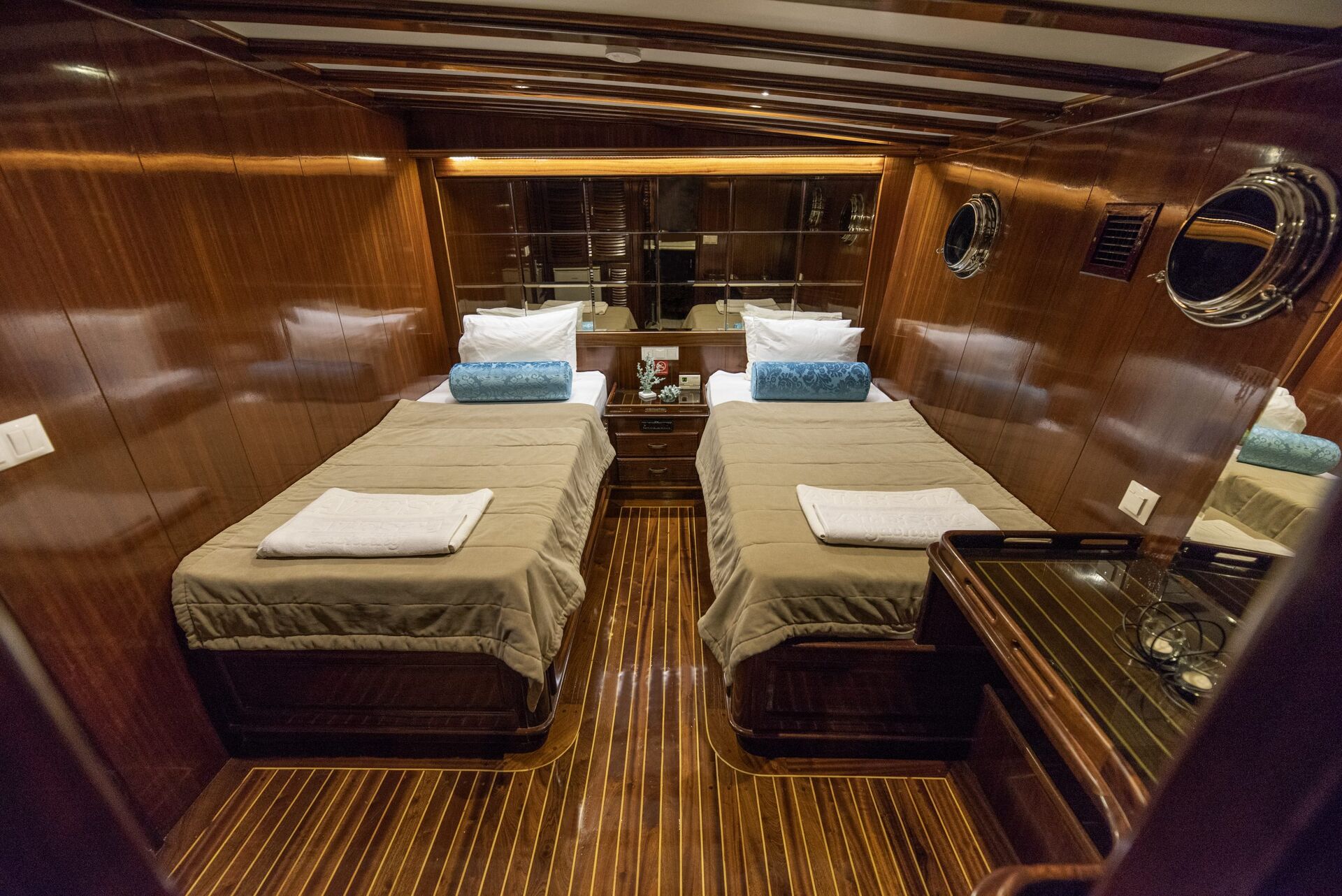 Luxurious yacht cabin with two single beds, beige bedding, blue accent pillows, wood-paneled walls, glossy wooden floor, porthole windows, and a nightstand with a telephone between the beds.