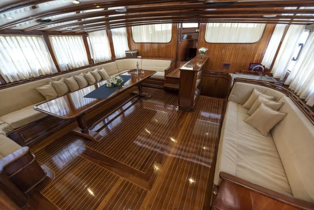 Luxurious yacht interior with polished wooden floors, cream-colored sofas with cushions, a large table with a floral centerpiece, and windows with sheer curtains, creating an elegant and spacious lounge area.