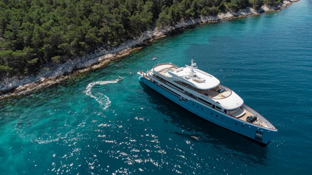 A large white yacht is anchored near a rocky, tree-covered shoreline in clear blue water. A small motorboat creates ripples as it circles close to the yacht. The scene is bathed in sunlight.