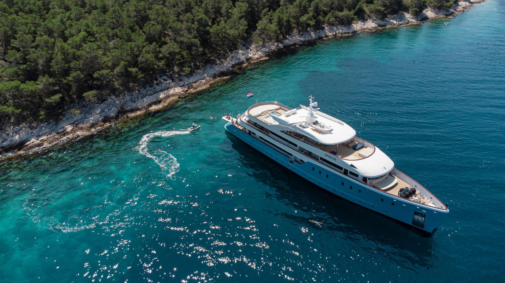 A large white yacht is anchored near a rocky, tree-covered shoreline in clear blue water. A small motorboat creates ripples as it circles close to the yacht. The scene is bathed in sunlight.