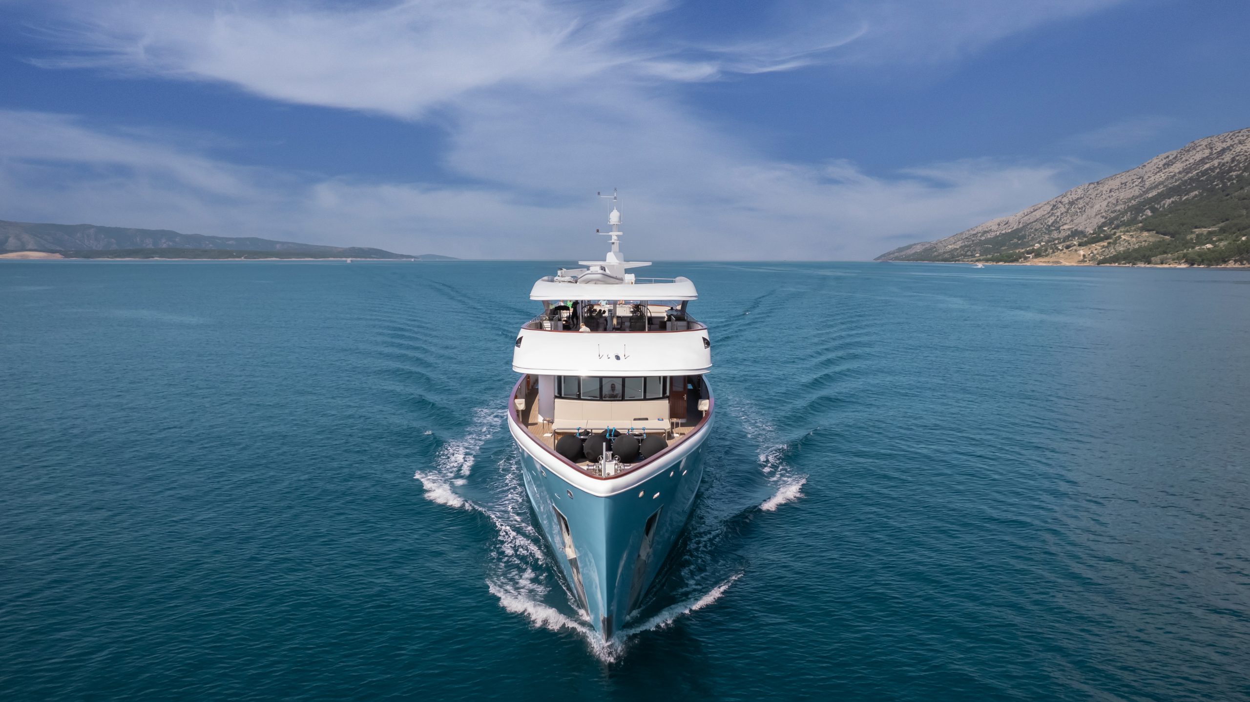 A large yacht sails through calm, blue waters with mountains visible on the right and a partly cloudy sky above.