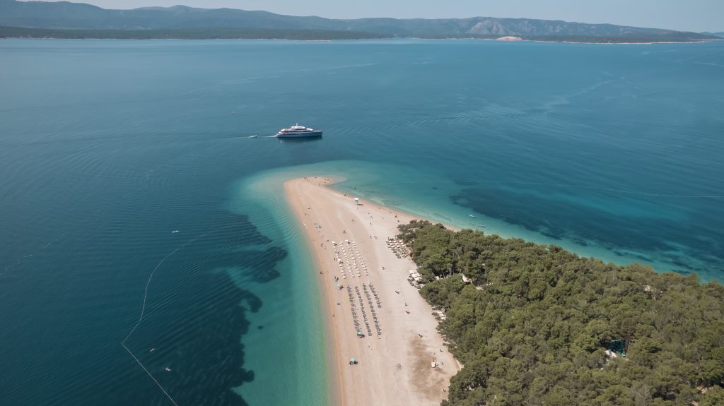 Aerial view of a sandy beach with rows of umbrellas and lounge chairs, surrounded by turquoise water and dense green trees. A white boat floats nearby, and distant mountains are visible across the sea.