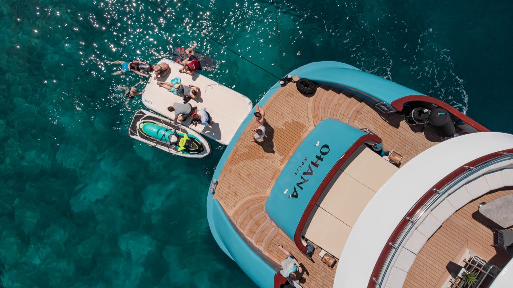 Aerial view of people relaxing on a yacht and a floating platform in clear blue water; some are sunbathing, others interact near a jet ski, with the yachts name OHANA visible on the deck.