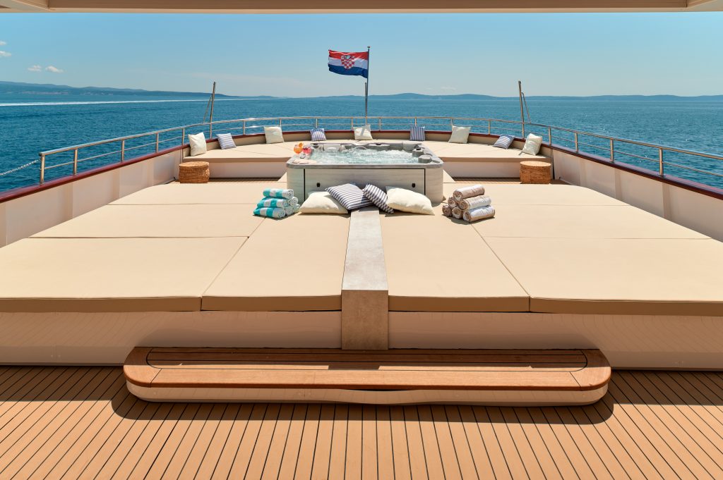 A luxury yacht deck features sun loungers, a hot tub, towels, and pillows, with a Croatian flag at the back. The yacht is on calm blue water under a clear sky, with distant land visible on the horizon.