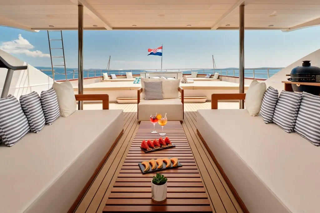 A luxurious yacht deck with beige cushioned seating, striped pillows, a wooden table with fruit and drinks, and a view of the sea. A Croatian flag waves at the stern under a clear blue sky.