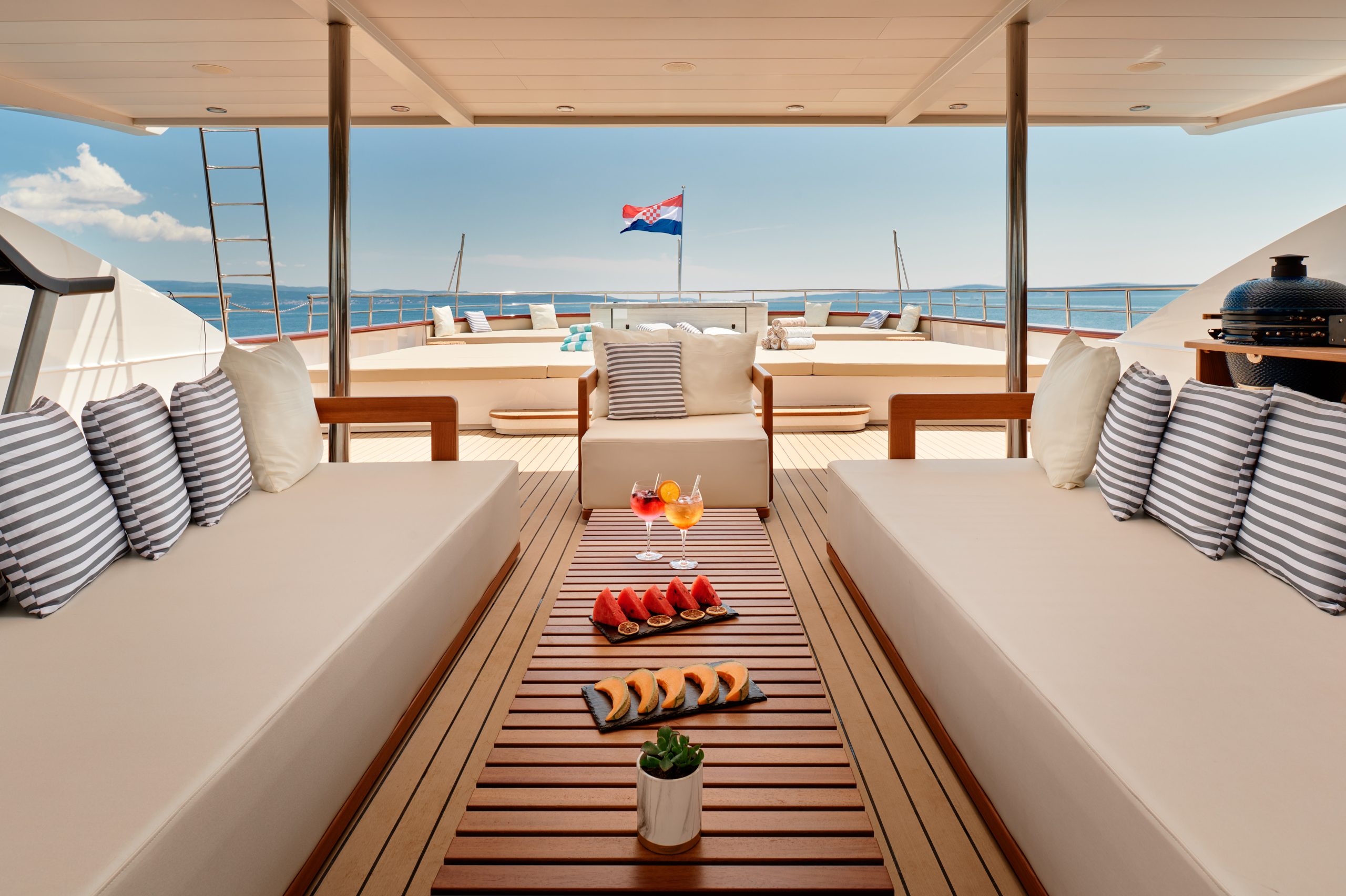 A luxurious yacht deck with beige cushioned seating, striped pillows, a wooden table with fruit and drinks, and a view of the sea. A Croatian flag waves at the stern under a clear blue sky.