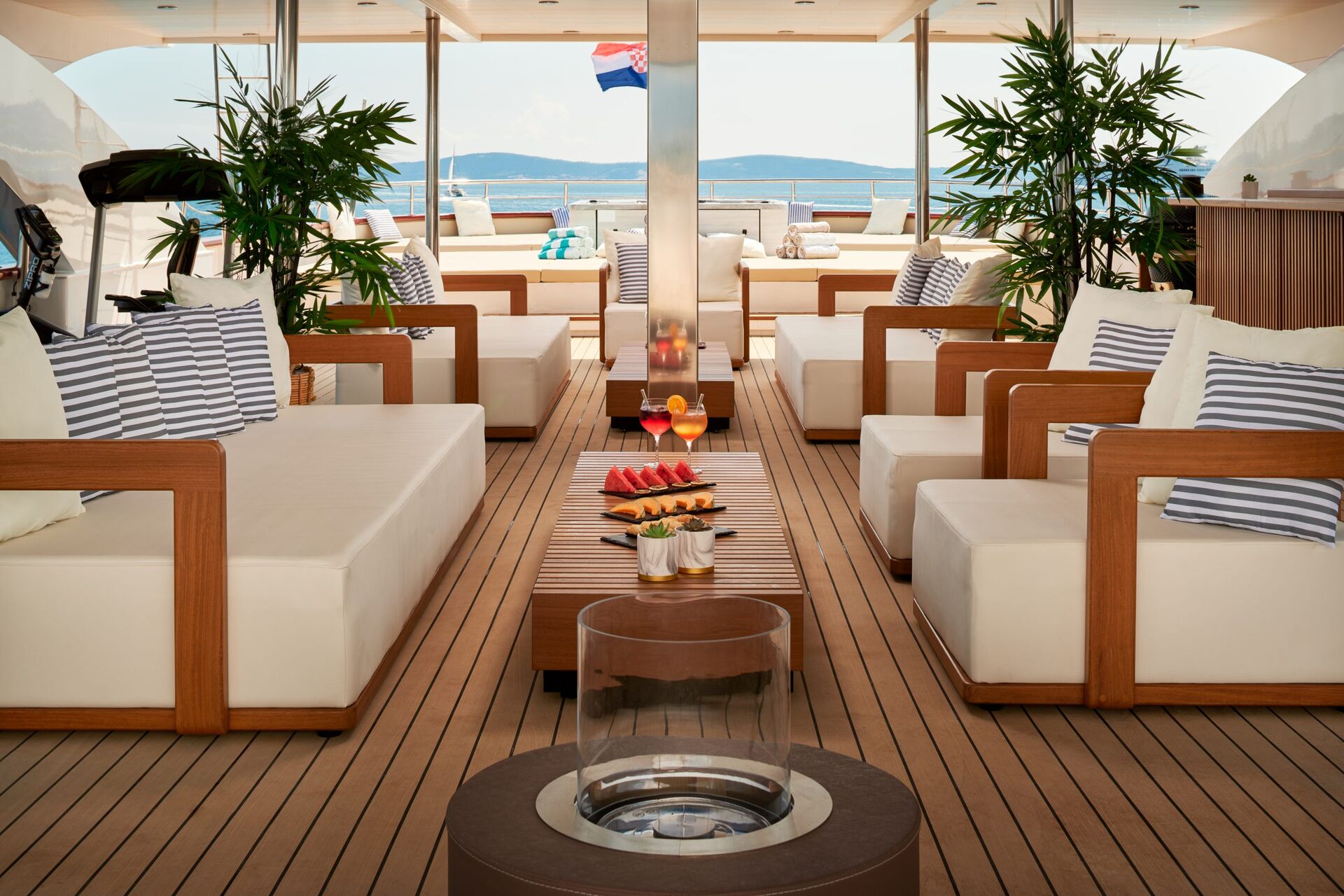 Luxurious yacht deck with white cushioned wooden seating, striped pillows, drinks, and a plate of watermelon. Plants decorate the sides and an ocean view with a Dutch flag is visible in the background.