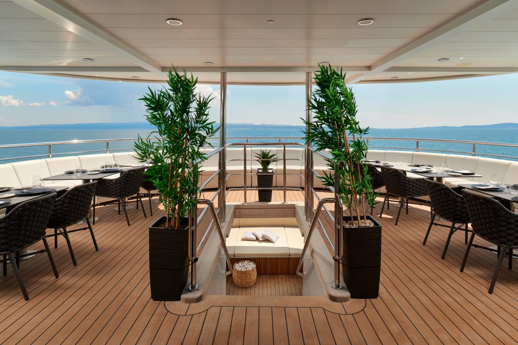 A luxurious yacht deck with wooden flooring, circular seating areas, potted plants, and a sunken lounging space at the center. The deck overlooks calm blue ocean waters under a partly cloudy sky.