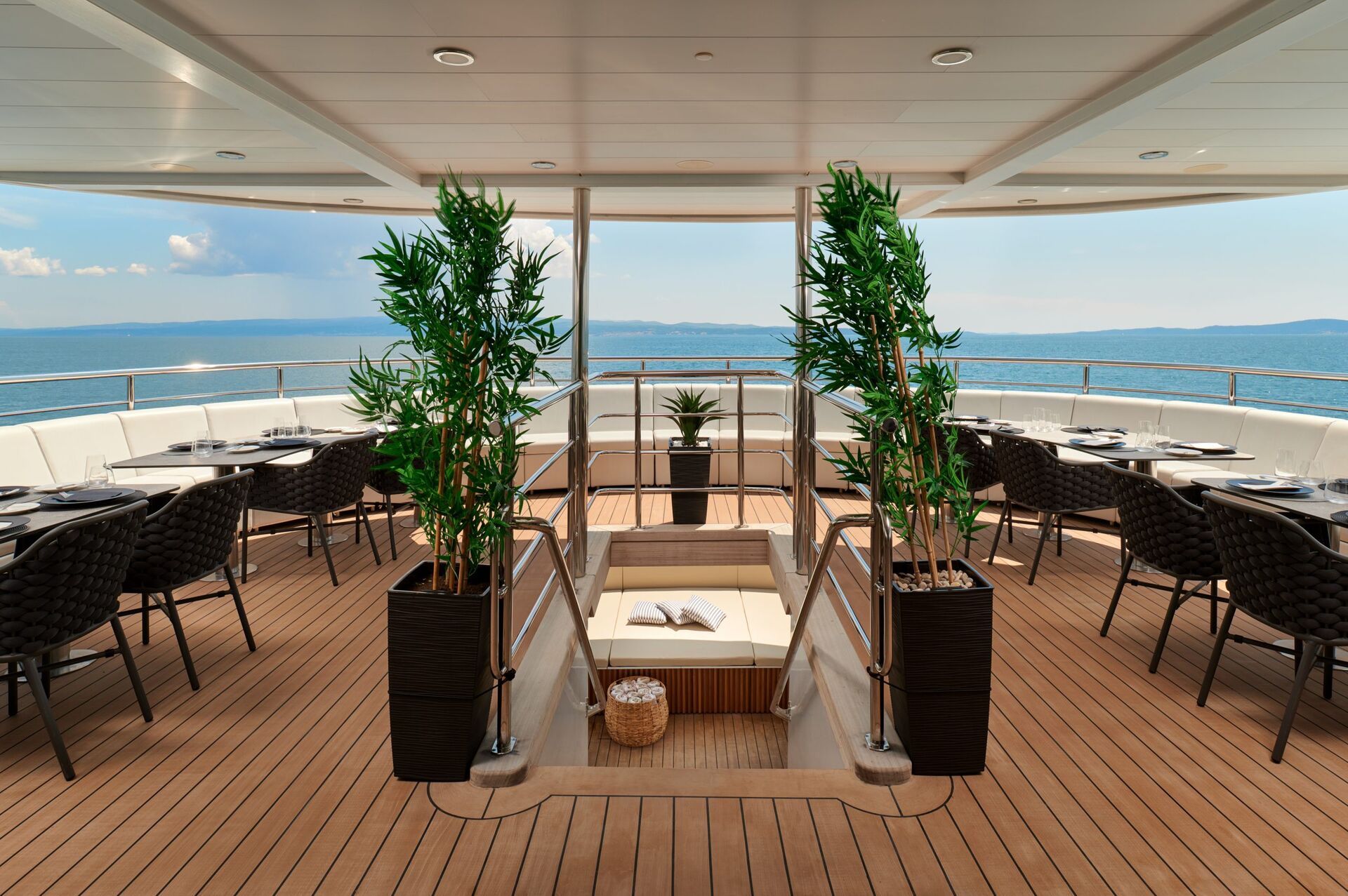 A luxurious yacht deck with wooden flooring, circular seating areas, potted plants, and a sunken lounging space at the center. The deck overlooks calm blue ocean waters under a partly cloudy sky.