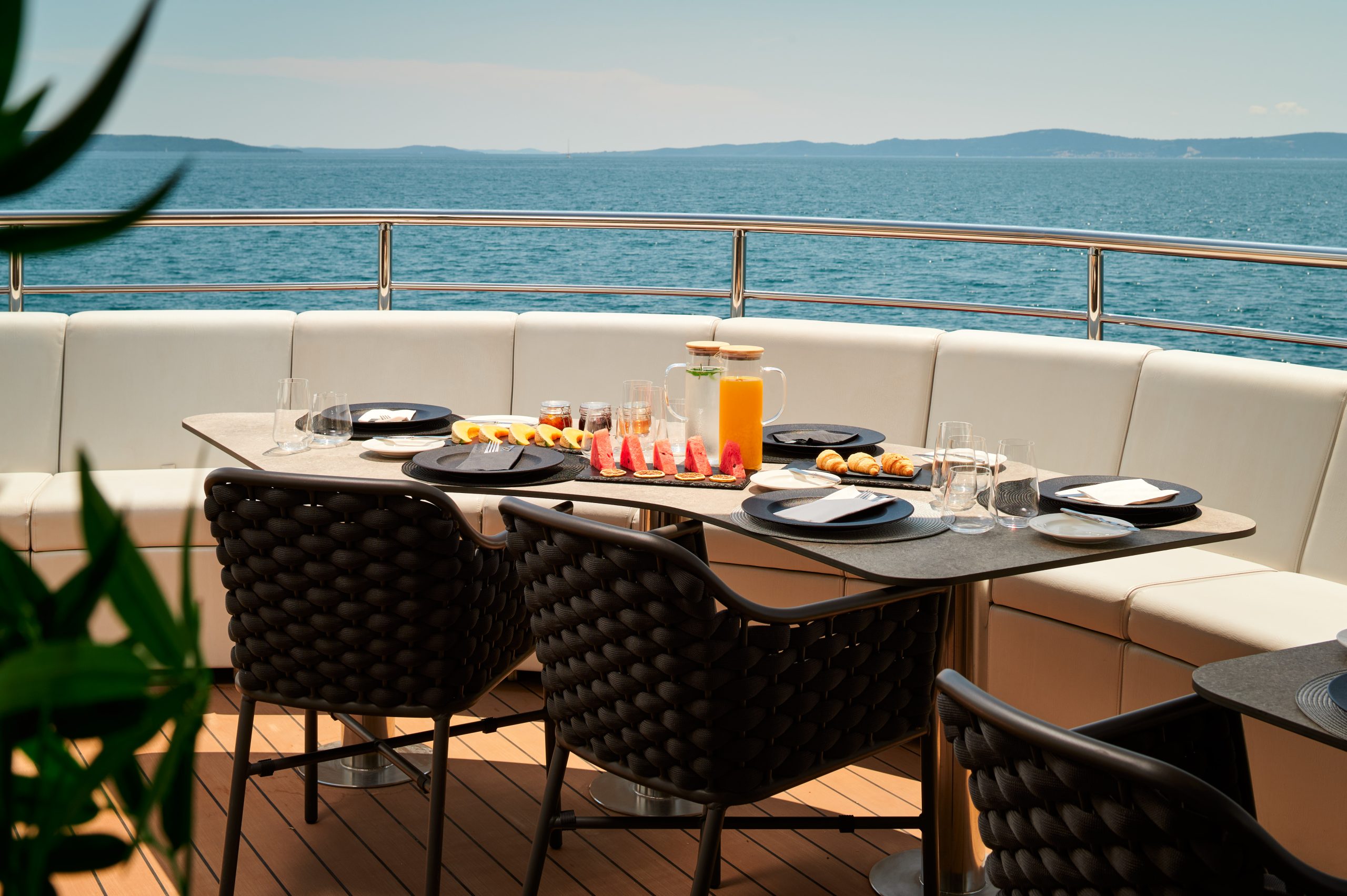 A table set for breakfast with juice, fruit, and pastries on a yacht deck, surrounded by cushioned seating with a view of the sea and distant mountains under a clear sky.