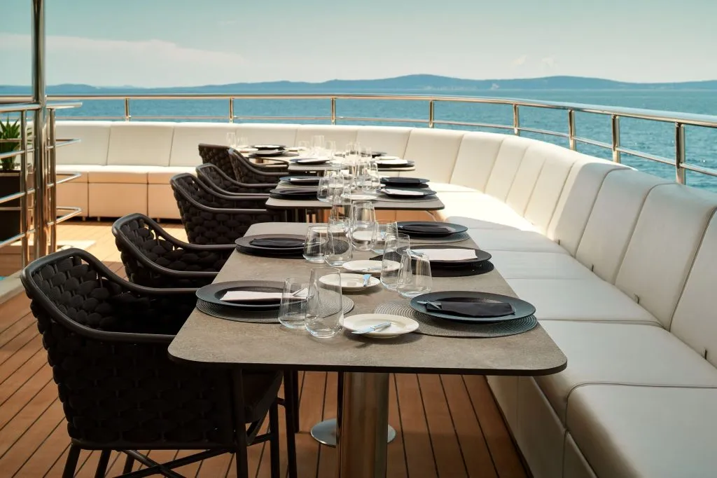 An elegant outdoor dining area on a yacht, featuring a long table set with black plates, glasses, and white napkins, surrounded by cushioned chairs and benches, overlooking the sea and distant hills.