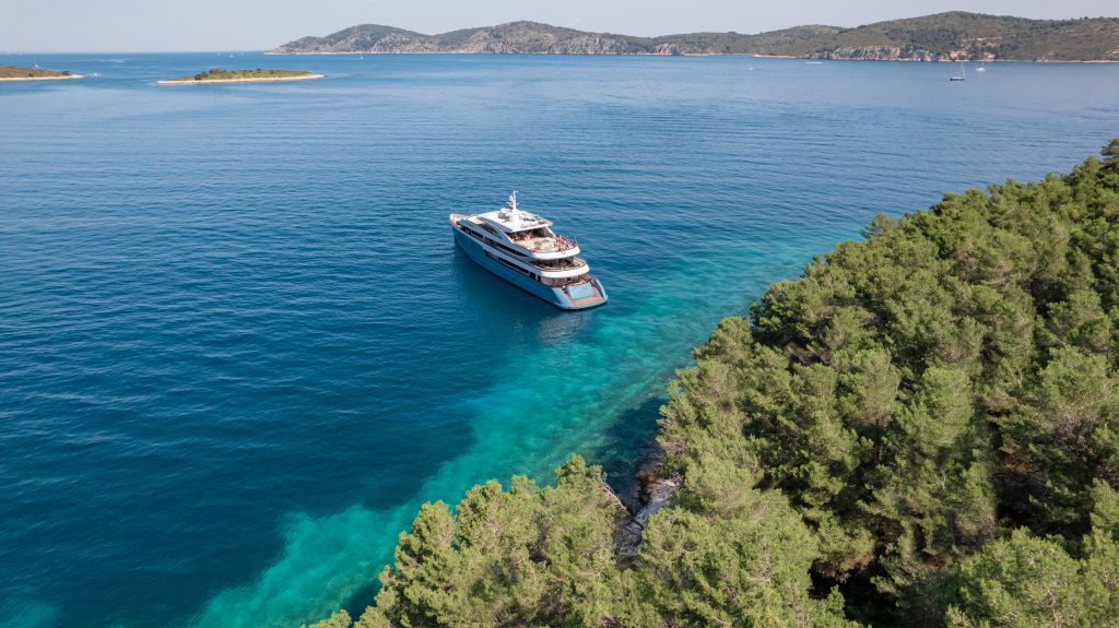 A large white yacht floats on clear blue water near a forested shoreline, with tree-covered islands visible in the distance under a clear sky.