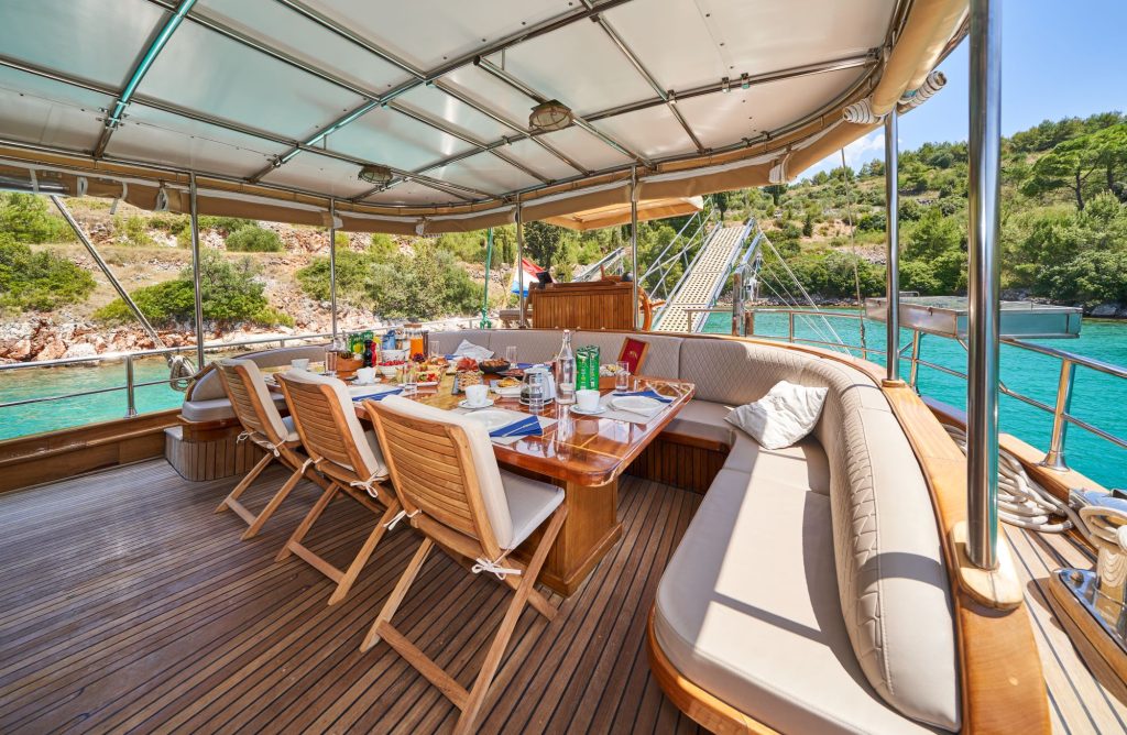 LIBRA A dining area on a yacht features a wooden table set for a meal with drinks and food. Cushioned benches and chairs surround the table, and turquoise water and a green, tree-covered coast are visible in the background.