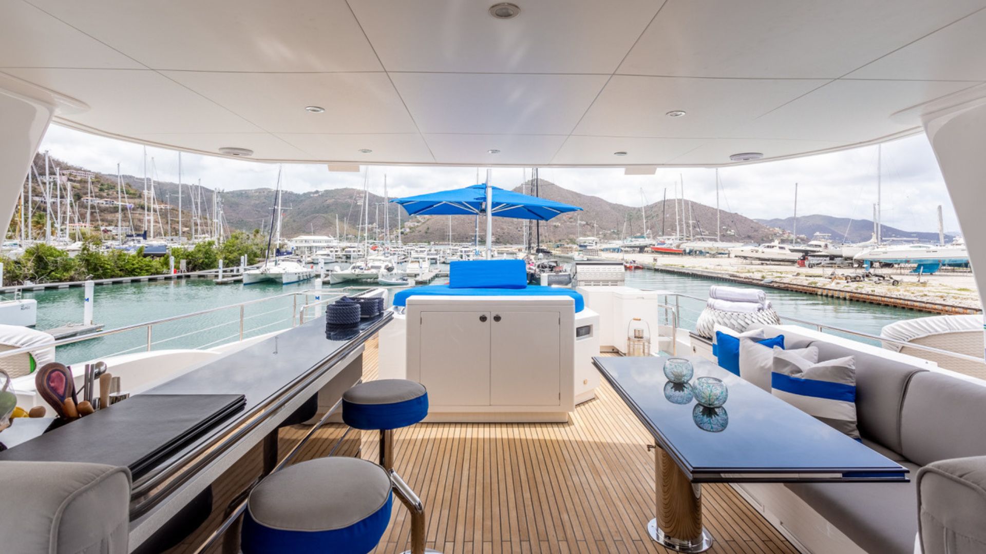 The image shows the deck of a luxury yacht with stylish seating, blue cushions, a table, and an umbrella. The yacht is docked at a marina with sailboats and hills visible in the background under a bright sky.