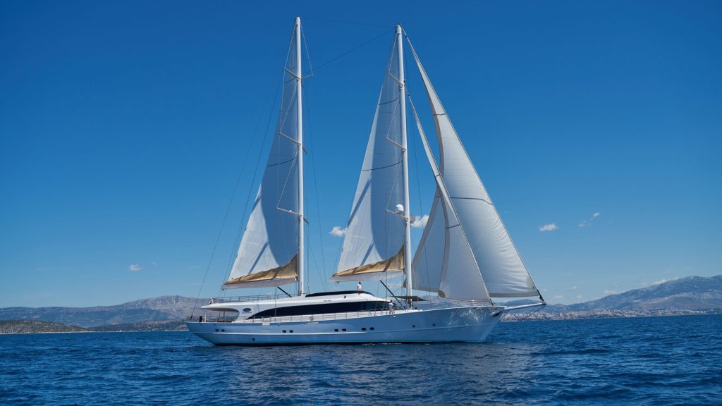 A large white sailing yacht with two tall masts and fully raised sails glides on calm blue water under a clear sky, with distant mountains visible on the horizon.