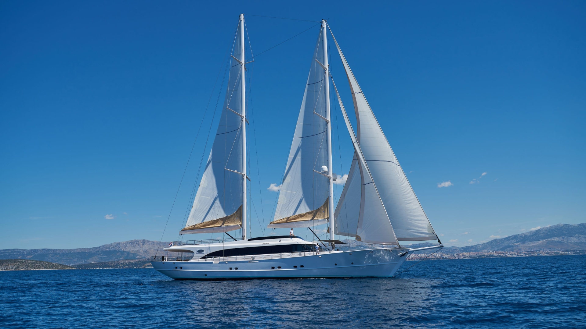 A large white sailing yacht with two tall masts and fully raised sails glides on calm blue water under a clear sky, with distant mountains visible on the horizon.