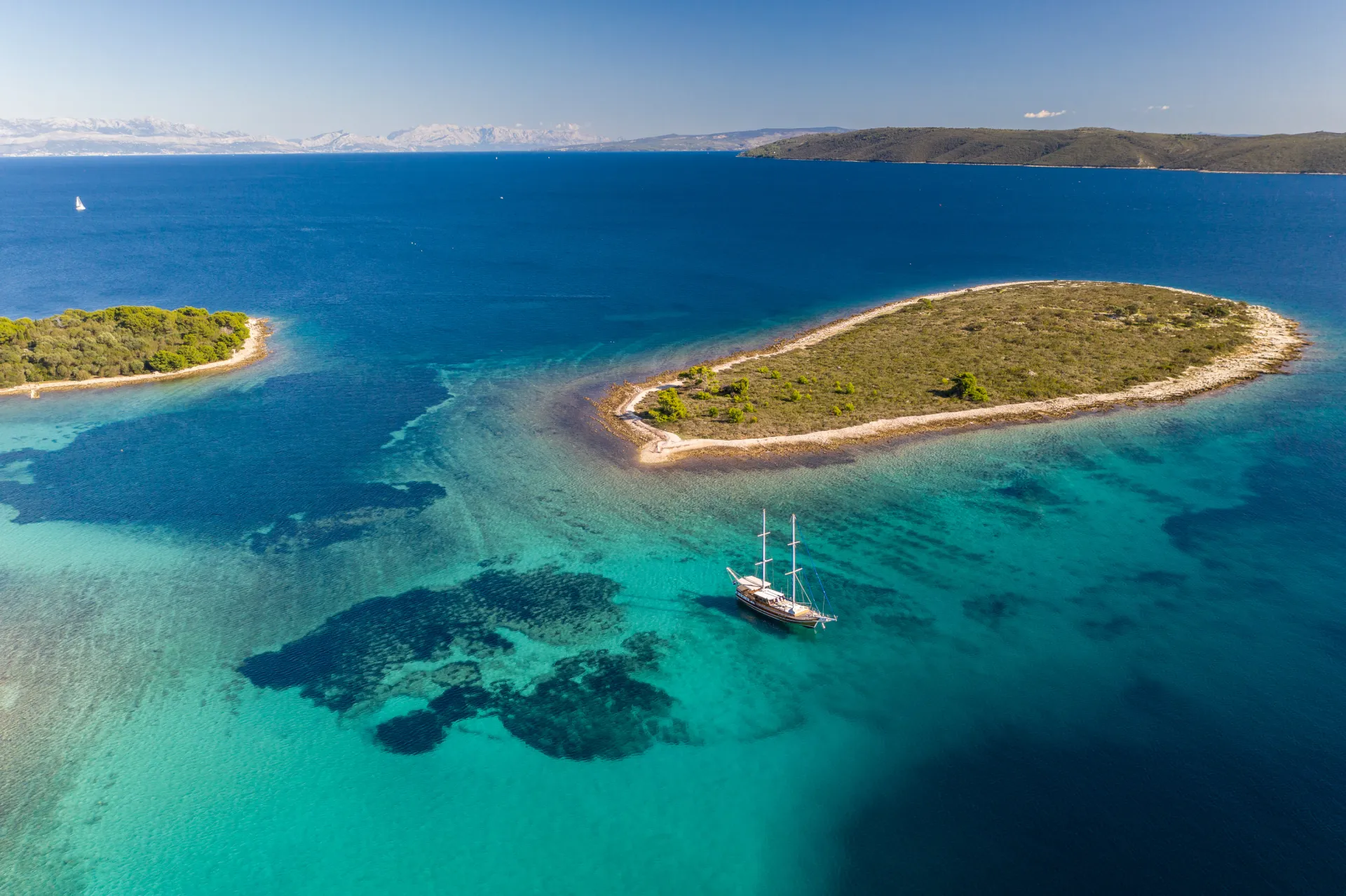 ANDI STAR Aerial view of a sailboat anchored near two small, lush islands surrounded by clear turquoise and deep blue sea under a bright, sunny sky.