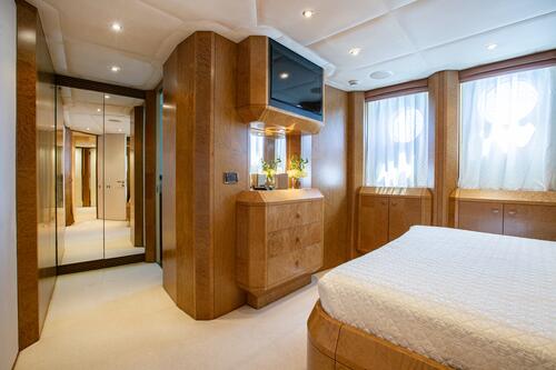 A modern yacht bedroom with wood-paneled walls, a bed covered in a white patterned bedspread, a wall-mounted TV above a dresser, two windows with sheer curtains, and mirrored closet doors.