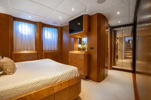 A luxurious yacht bedroom with wood-paneled walls, a large bed with a white quilt, two round windows with curtains, built-in cabinets, a mounted TV, and a mirrored closet door reflecting a hallway.