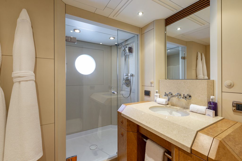 Modern bathroom with beige walls, a glass-enclosed shower featuring a round porthole window, a vanity with a sink, a large mirror, and two white bathrobes hanging on the wall.