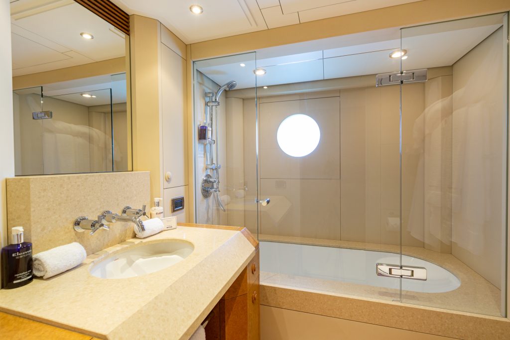 Modern bathroom with a beige countertop, sink, and faucet, plus toiletries and towels. A glass-enclosed bathtub and shower area features a round window and built-in shelf, with neutral walls and bright lighting.