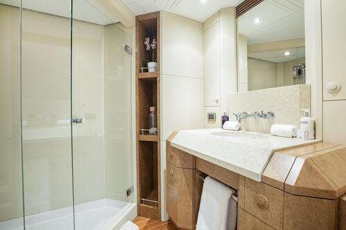 Modern bathroom with a glass-enclosed shower, light-colored cabinets, wooden shelves with toiletries, a large mirror, and a countertop with a sink, two rolled towels, and various bath products.
