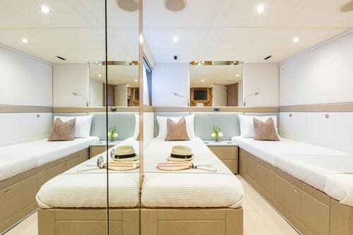 A modern, well-lit cabin with two single beds, beige bedding, and decorative pillows. Mirrors on the walls create a spacious feel. A hat, sunglasses, and tray sit on one bed; small plants and lamps decorate the space.