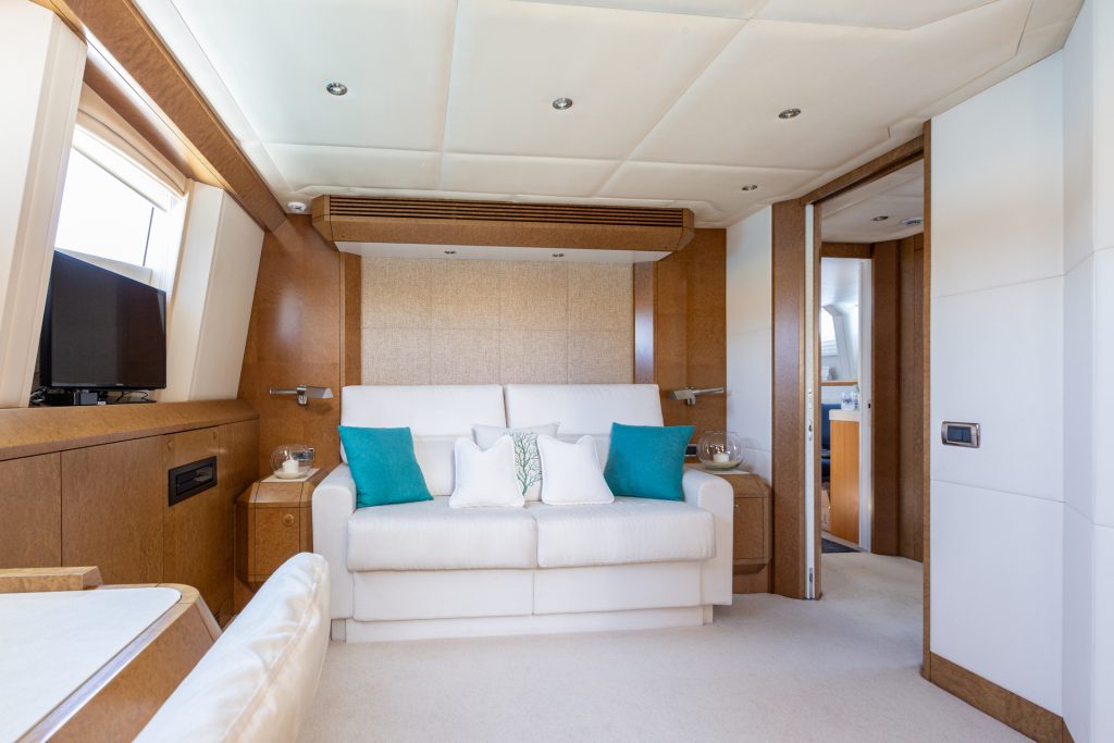 A modern yacht interior with a white sofa, teal and white pillows, wood paneling, light beige carpet, and large windows letting in natural light. There is a doorway leading to another room.