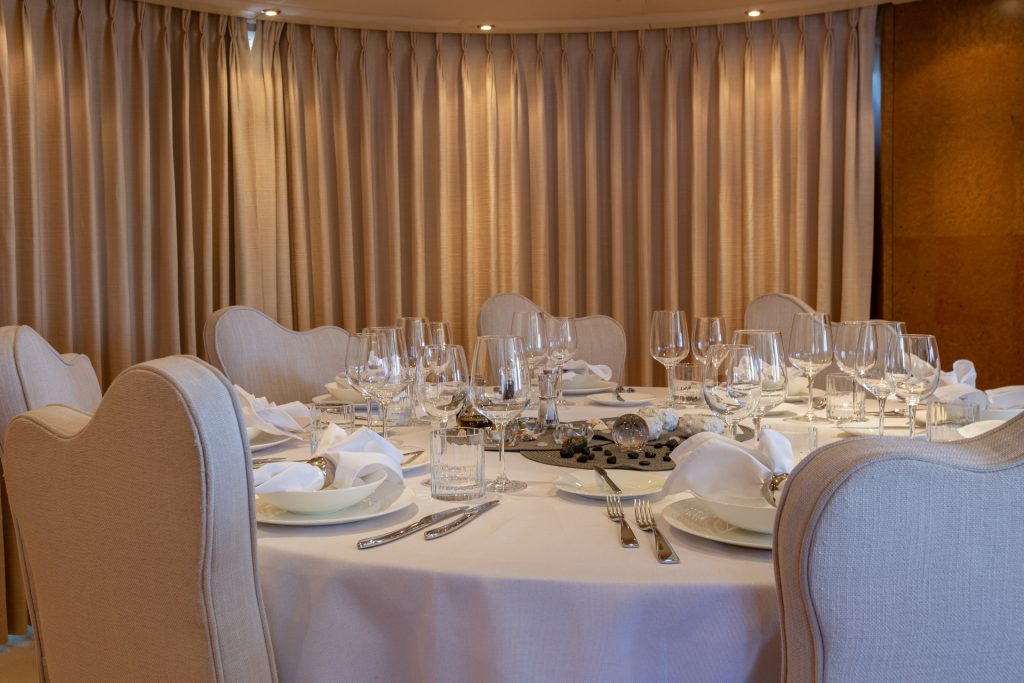 A round dining table set for six with white linen, neatly folded napkins, glassware, and silverware, surrounded by upholstered chairs in a room with beige curtains and soft lighting.