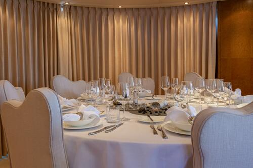 A round dining table set for six with white linen, neatly folded napkins, glassware, and silverware, surrounded by upholstered chairs in a room with beige curtains and soft lighting.