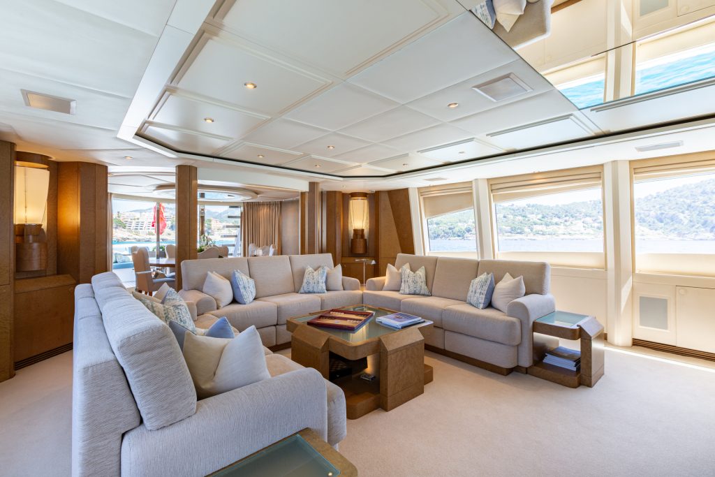 A spacious yacht lounge with large windows, beige sofas, decorative pillows, and a central wooden coffee table, offering views of the sea and coastline outside. The room is bright with modern, elegant decor.