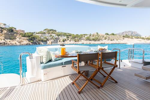 BENITA BLUE A table set with drinks and food sits on the deck of a yacht, overlooking clear blue water and rocky coastal cliffs with houses under a sunny, clear sky.