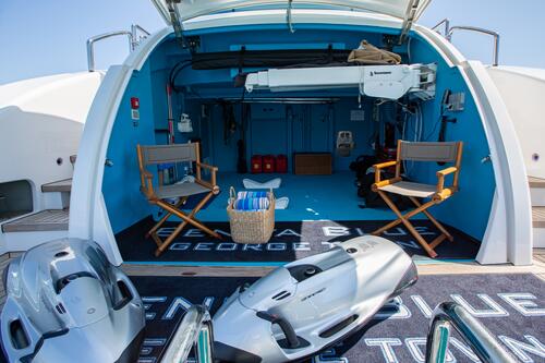 The open back area of a yacht with two director’s chairs, a basket with towels, and water sports equipment. The space is clean, organized, and painted blue, with natural light and ocean visible in the background.