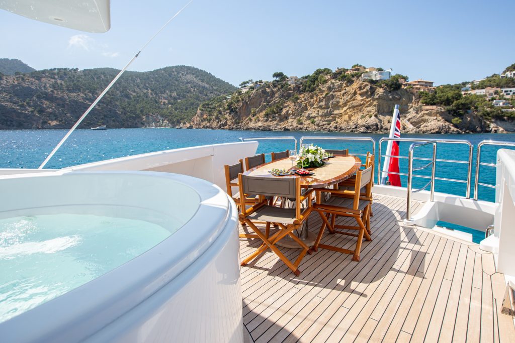 A yacht deck features a hot tub and a dining table with chairs, set with flowers and refreshments. The yacht is on clear blue water, with rocky hills and houses visible in the background under a sunny sky.