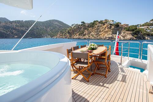 A yacht deck features a hot tub and a dining table with chairs, set with flowers and refreshments. The yacht is on clear blue water, with rocky hills and houses visible in the background under a sunny sky.