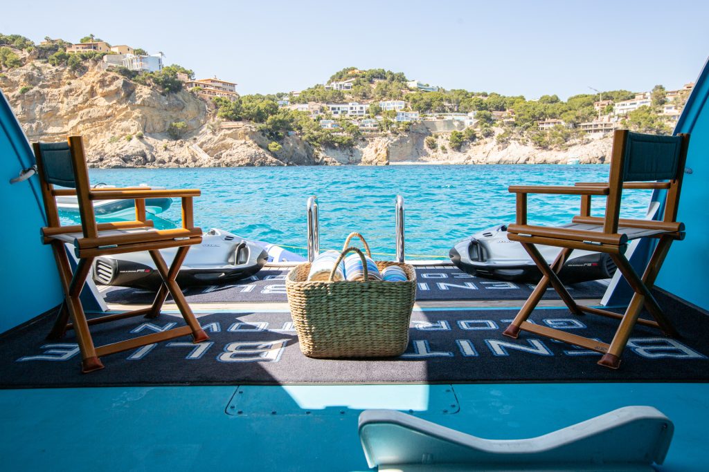 Two wooden chairs face the turquoise sea from a boat’s open deck, with a straw basket placed between them. Rocky cliffs and houses are visible across the water under a clear sky.