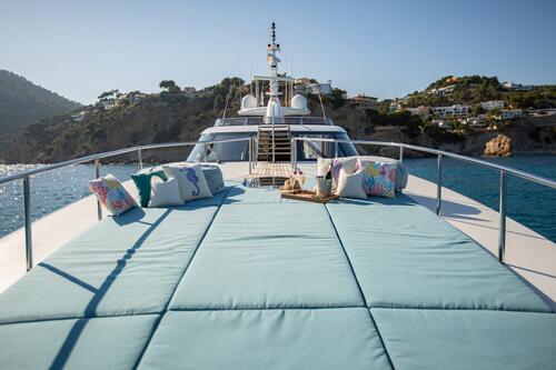 A large yacht deck with light blue cushioned loungers, decorative pillows, and a tray with drinks, overlooking clear blue water and coastal hills with houses in the background.