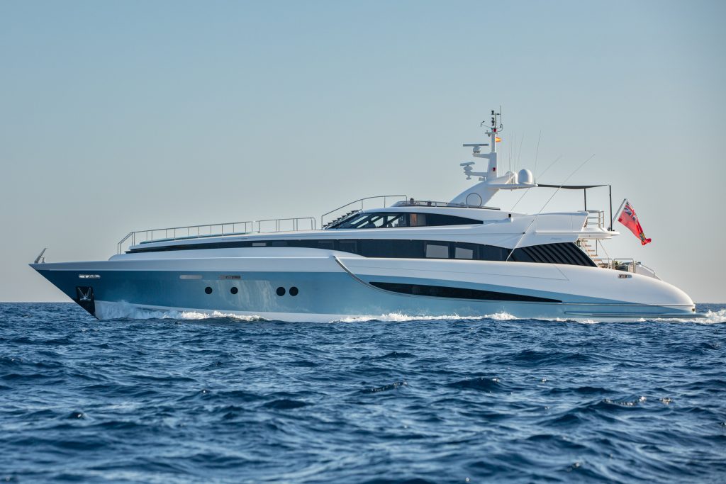 A large white luxury yacht with a sleek modern design cruises on calm blue ocean water under a clear sky. A red flag is visible at the stern of the yacht.