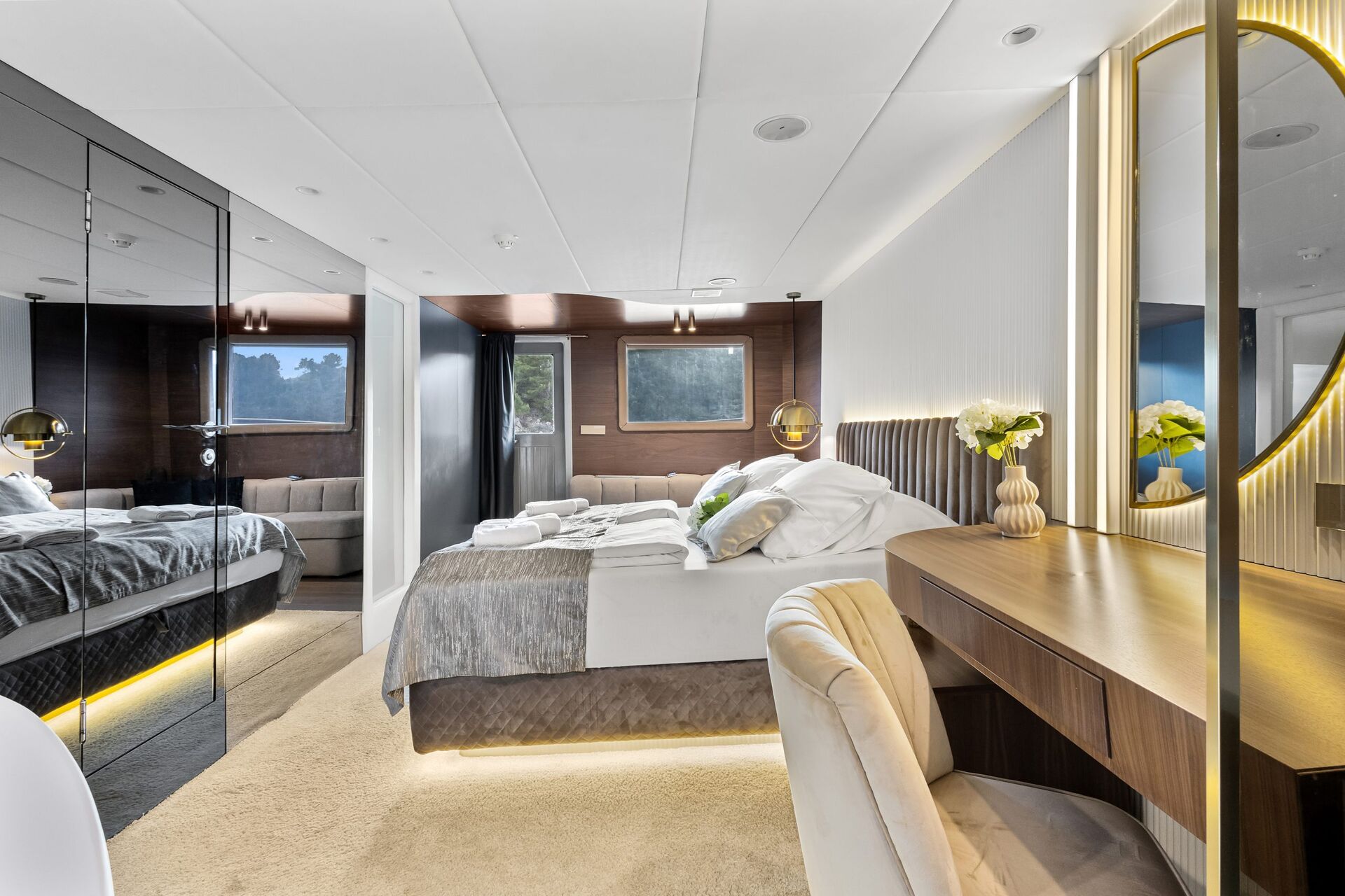 Modern, luxurious bedroom with a neatly made double bed, mirrored wardrobe, vanity table with a round lit mirror, and fresh flowers. Large windows and soft lighting create an inviting, elegant atmosphere.