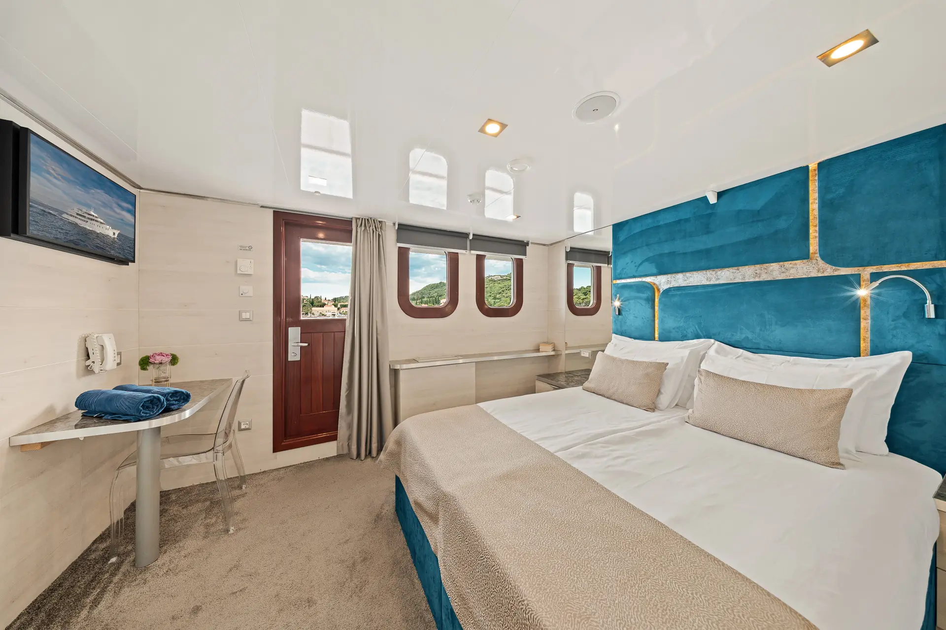 A modern cruise ship cabin with a large bed, blue and beige decor, three windows, a desk with a towel, wall-mounted TV, and a wooden door. Natural light fills the bright, spacious room.