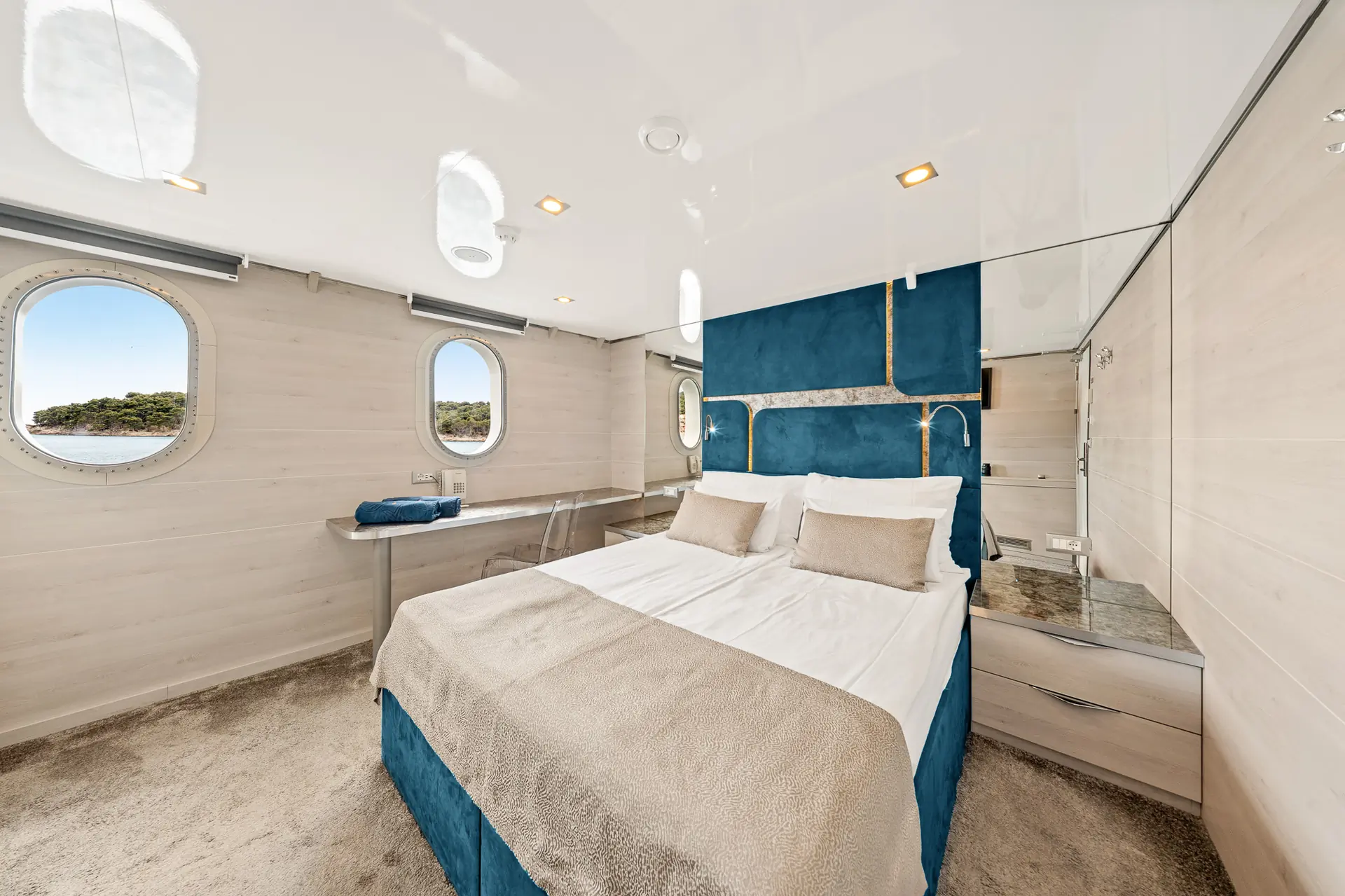 A bright, modern bedroom on a yacht with a double bed, blue and beige bedding, three round windows, a mirrored wall, and a view of the sea and greenery outside.