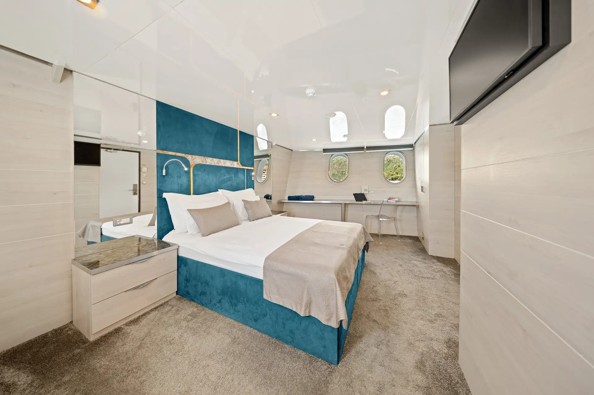 Modern bedroom with a double bed, teal and white decor, wall-mounted TV, mirrored surfaces, a desk with a clear chair, bedside tables, carpeted floor, and round porthole windows.