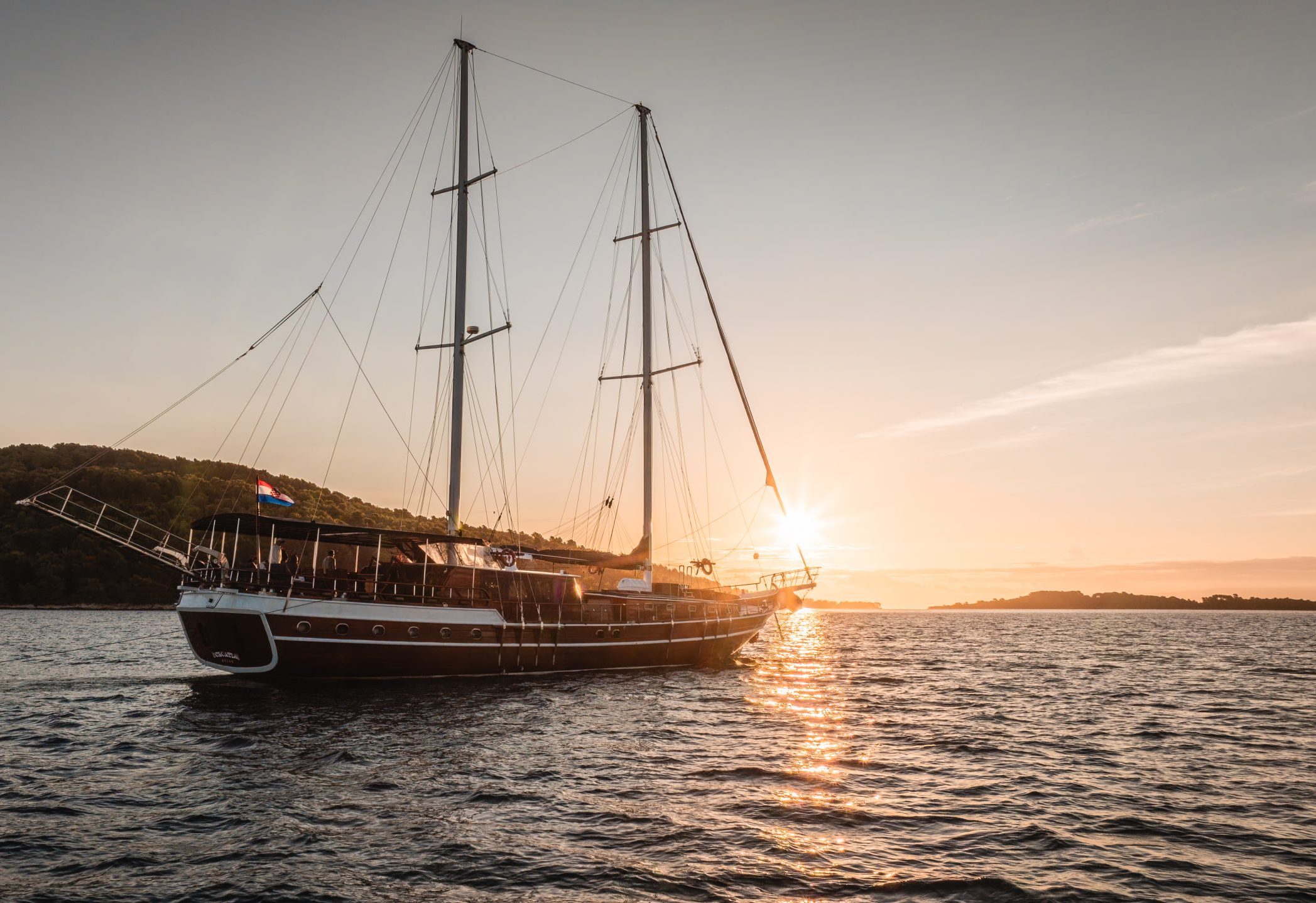 A large sailboat with tall masts sails on calm water at sunset, with the sun low on the horizon and a small island and wooded shoreline visible in the background.