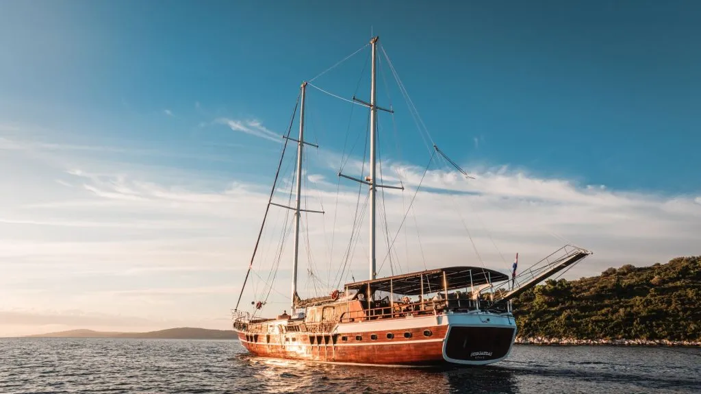 A large wooden sailboat with two tall masts floats on calm water near a green, hilly shoreline under a clear blue sky at sunset.